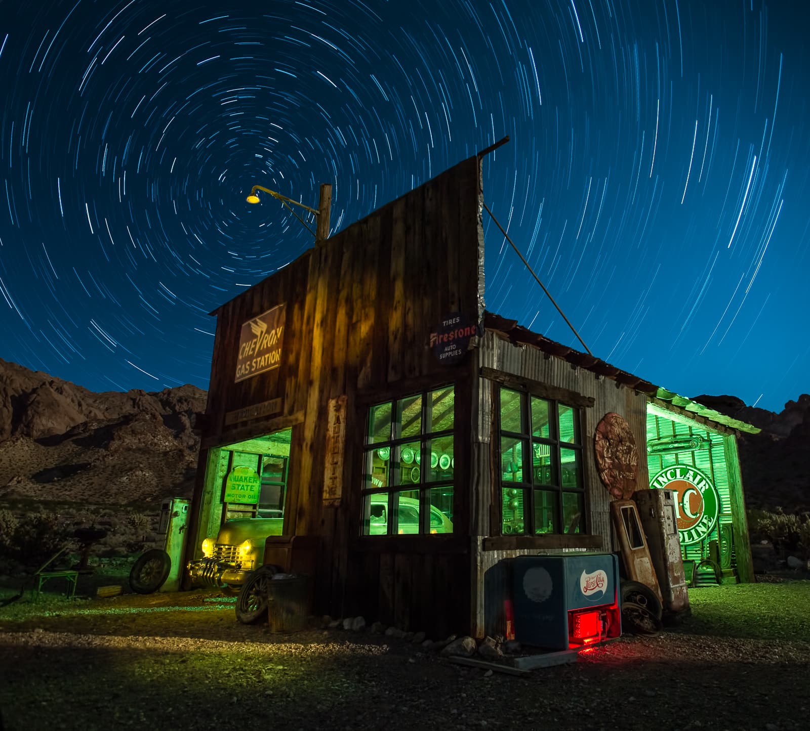 An old gas station lit green and gold beneath circular star trails